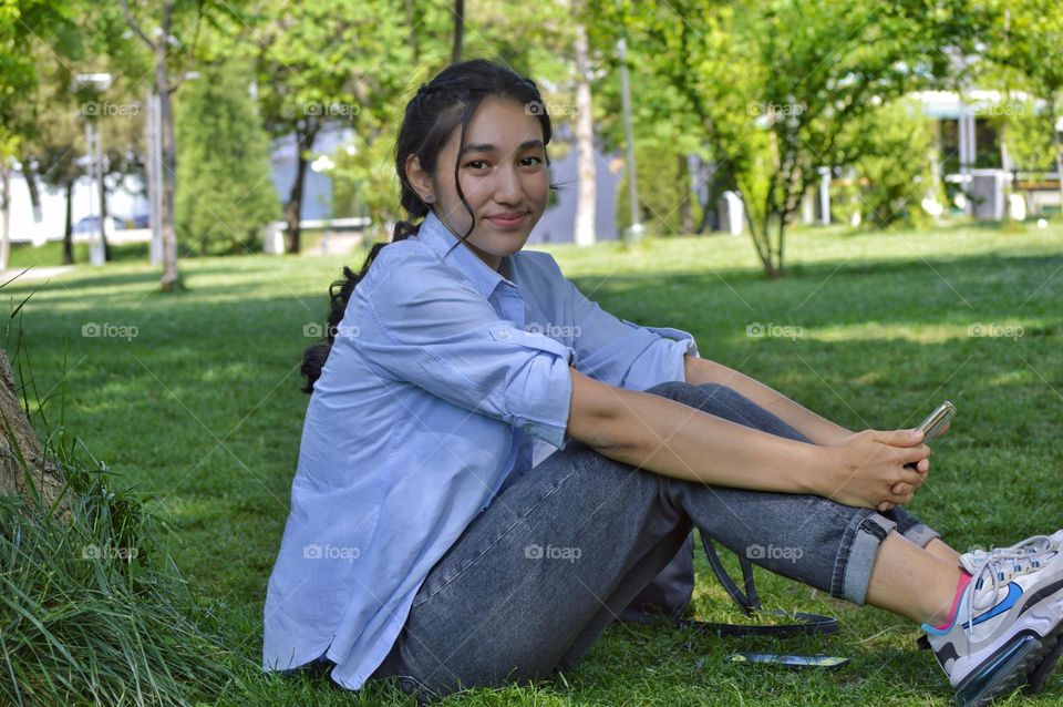 a girl in jeans and a shirt sits on a lawn in a park with a phone in her hands