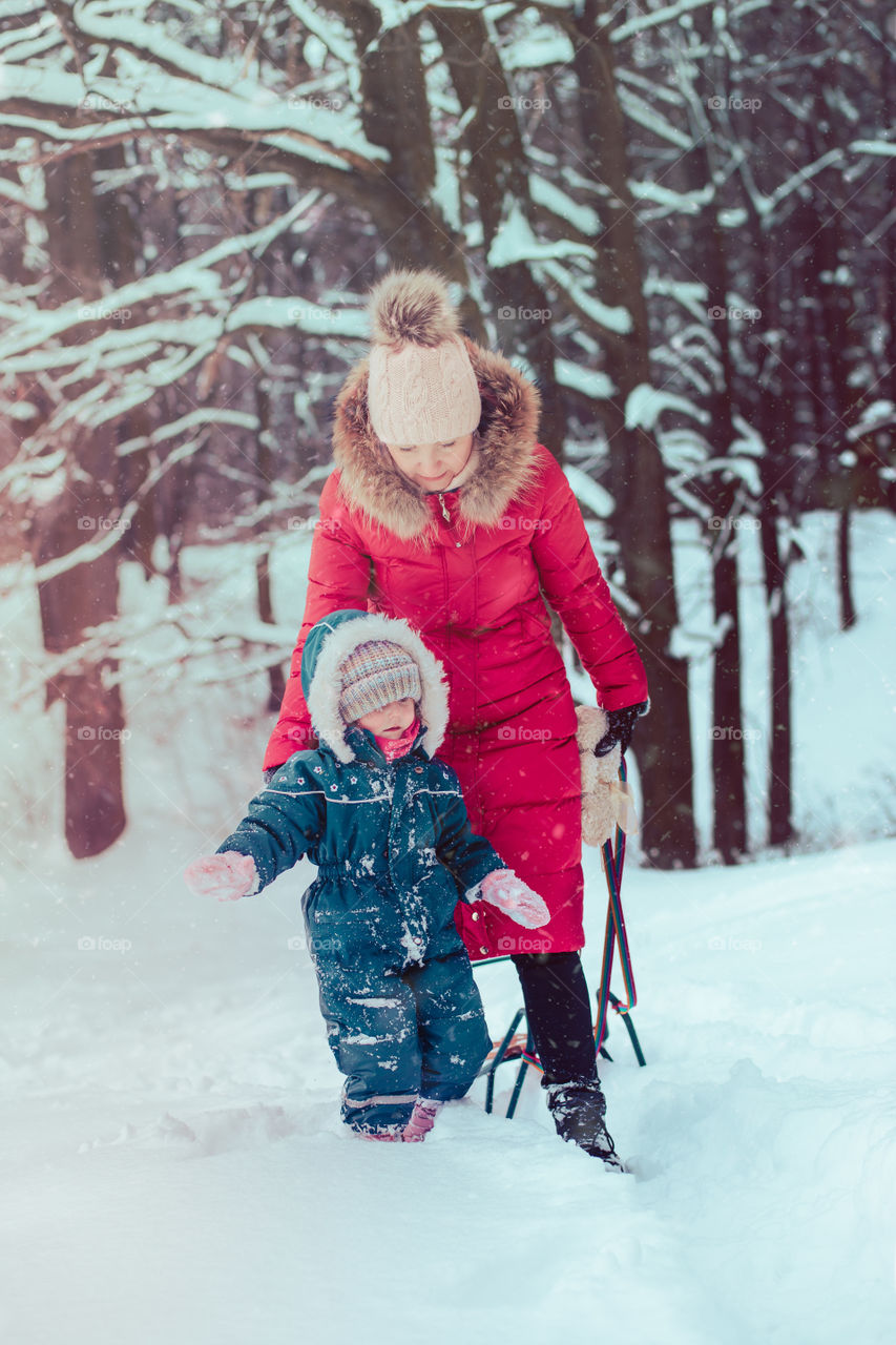 Mother and her little daughter are spending time together walking outdoors in forest in winter while snow falling. Woman is pulling sled, a few years old girl is walking through the deep snow, enjoying wintertime