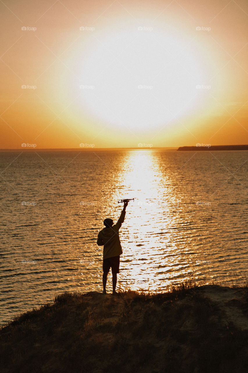 The silhouette of the man with a drone on coastline during sunset. Golden waves on the sea on background 