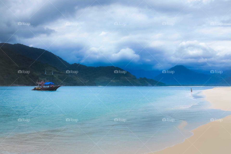 A woman looks at a ship, on the beach. There's a adventure sense, like the boat is rescuing her. Pirate Boat.