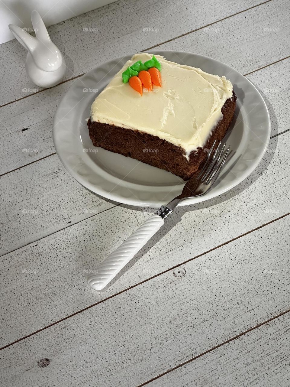 A slice of carrot cake with three little carrots decorating the icing on a white patterned plate with a white plastic swirl handled fork being overseen by a small white ceramic bunny, on a whitewashed wooden table