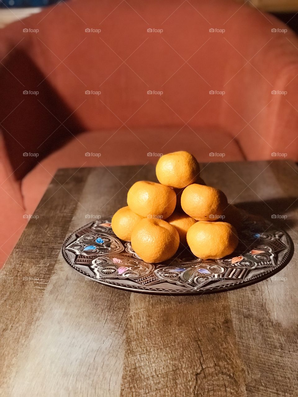 A portrait of a beautiful embellished metal plate of mandarin oranges. The plate is placed on a wood table and there is an orange chair behind the table.