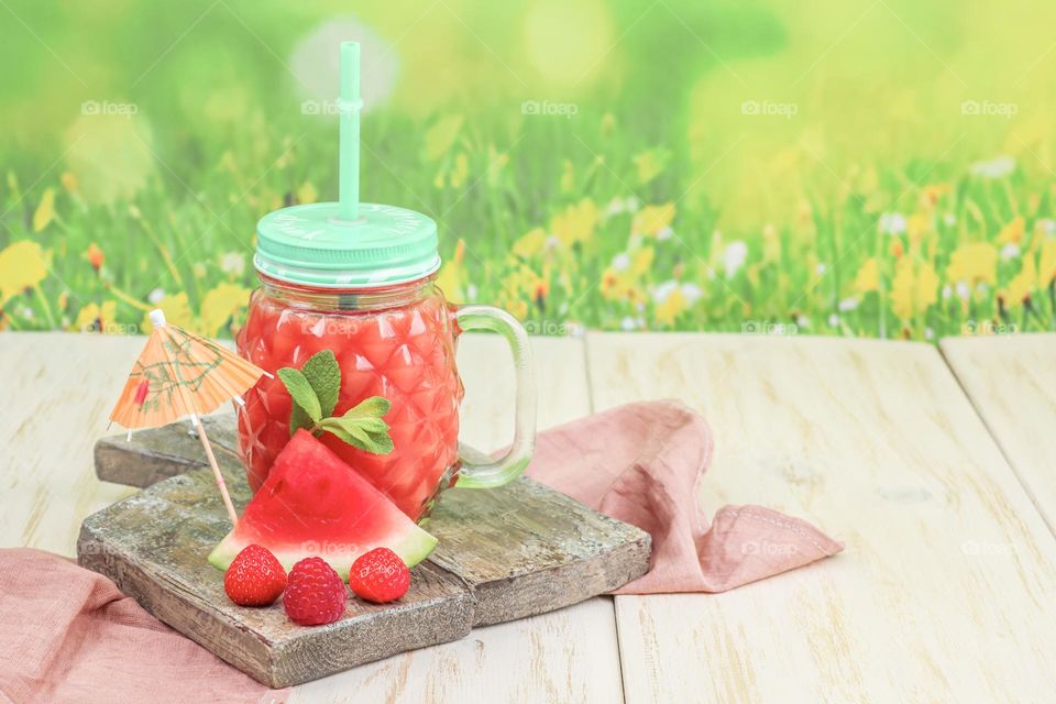 Watermelon-strawberry drink in a faceted jar with a handle, a metal lid and a straw stand on a wooden cutting board with a kitchen napkin from below with a piece of watermelon, a paper umbrella and fresh strawberries against a blurred green-yellow fi