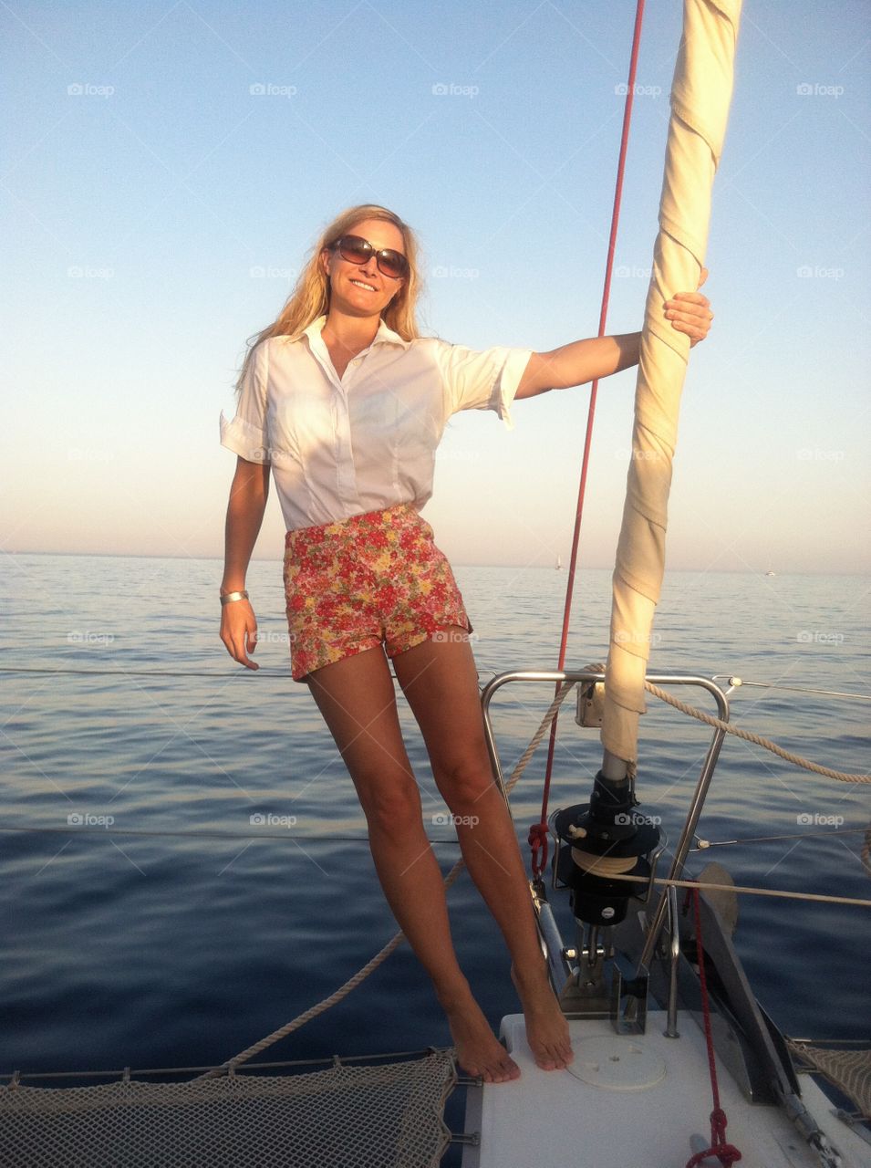 A young woman in colorful shorts hangs from the front sail of a small boat with the ocean behind her.  Santorini Island, Greece 2013.