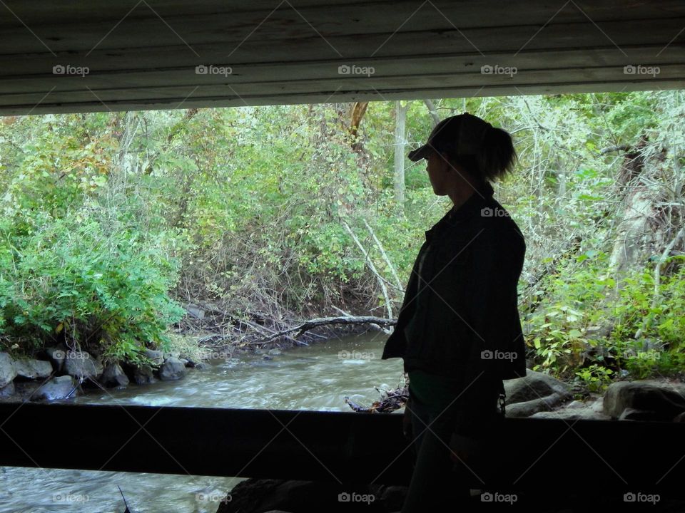 Woman standing under a bridge in a shadow near a Michigan river surrounded by green bushes, plants and trees. 