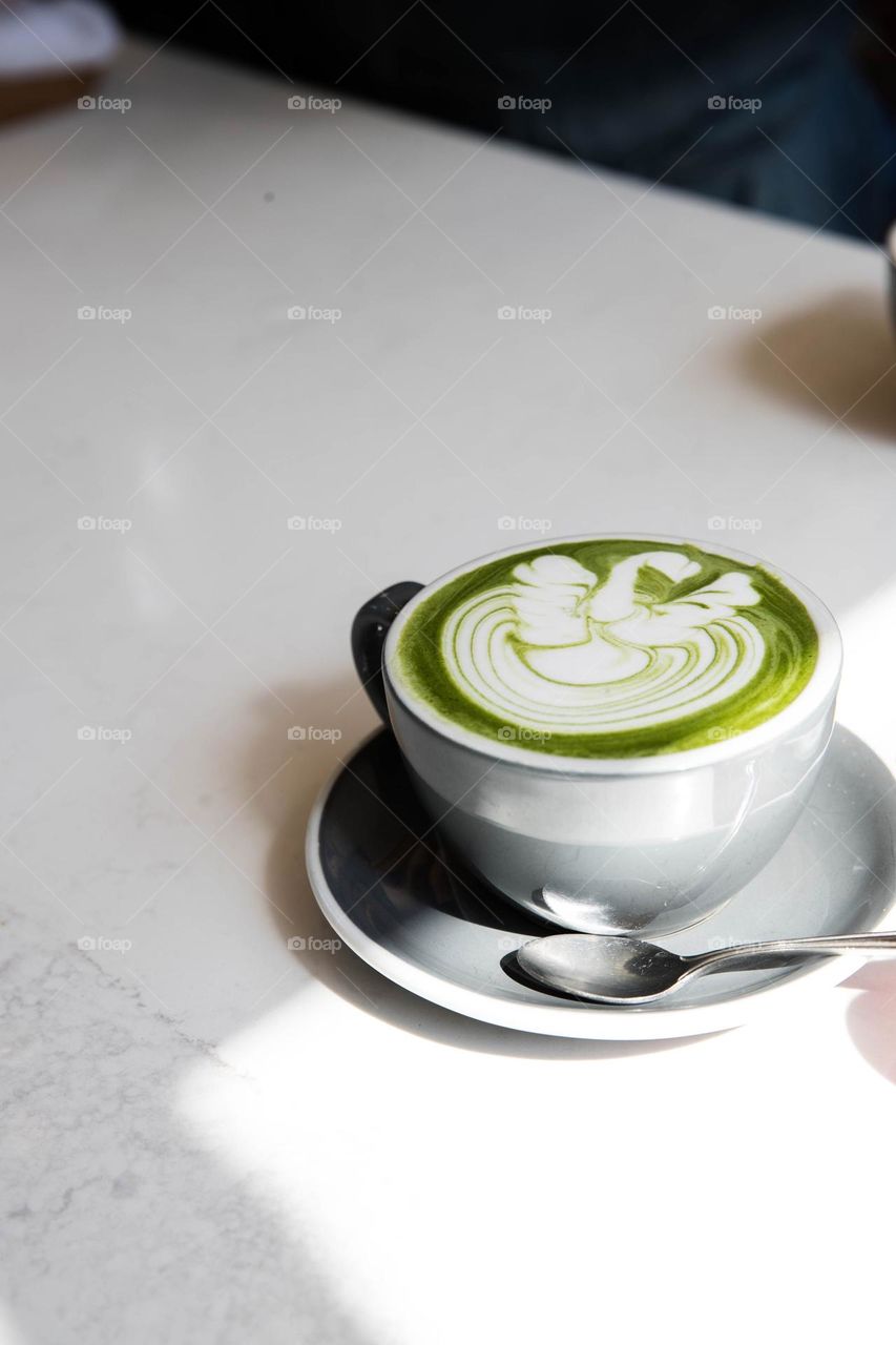 A gray cup and saucer with a silver spoon stands on a white table.
Inside the cup is a bright green matcha latte.