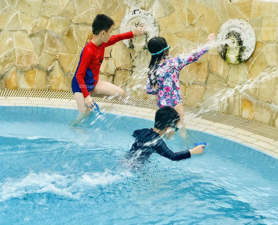 Children love to play and enjoy  at swimming pool in summer , here three children playing under flowing water (fountain) and water sprinklers in their hands . Totally it’s a fun time for everyone.