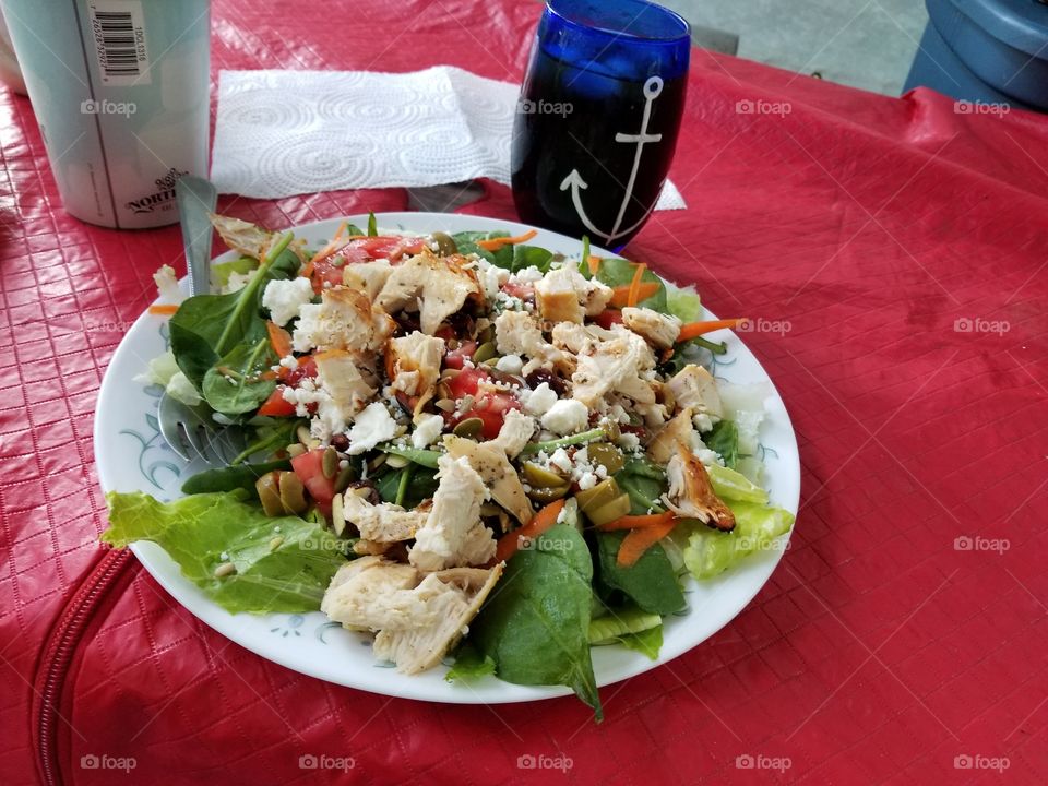 Chicken ceaser salad with chicken, lettuce, carrots, feta, and nuts with ranch dressing surrounded by a red tablecloth with a blue anchor glass.