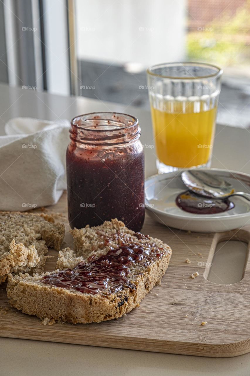The fresh morning light enters the kitchen where there is a slice of cereal bread and raisins barred with grape candy on a wooden board and orange juice 
