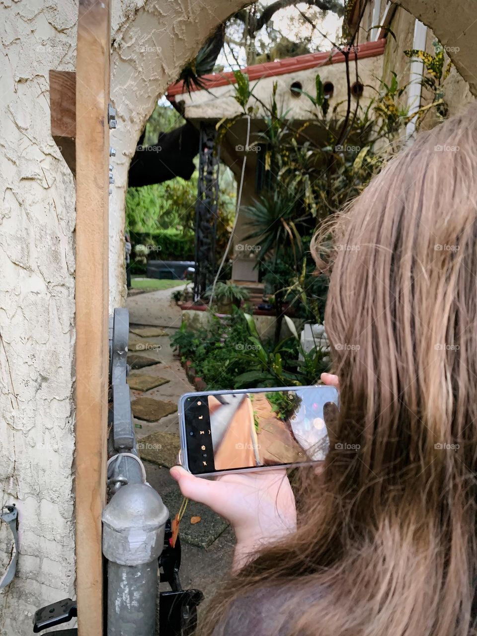 Spanish old style architecture residential large house built in the early 1900s. Backyard doorway with antique style, view on both side of the wall, young girl taking photos facing the tiled walkway sidewalk with trees in a beautiful tropical garden.