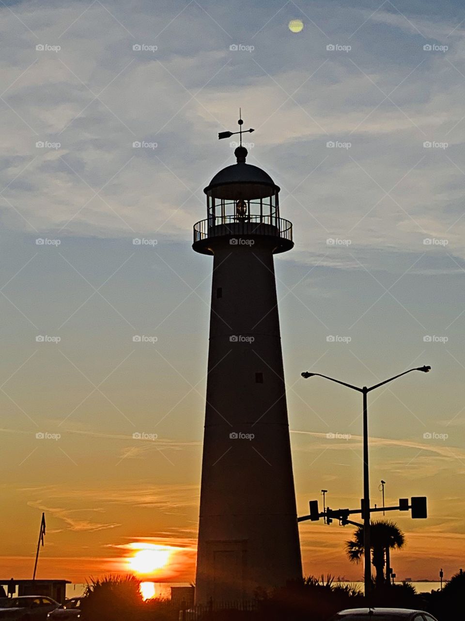 A silhouette of a lighthouse stands erect as the brilliant sunset and sparkling moon appear in the forefront 