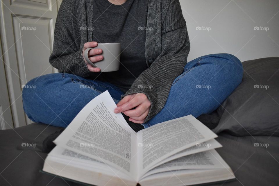 Woman sitting on the bed, while drinking coffee and reading a book. Holding a coffee mug or cup. Woman studying. Wearing denim jeans and grey jersey. White walls, grey and blue clothing. Hand, legs and fingers. Grey T-shirt and comfortable jeans.