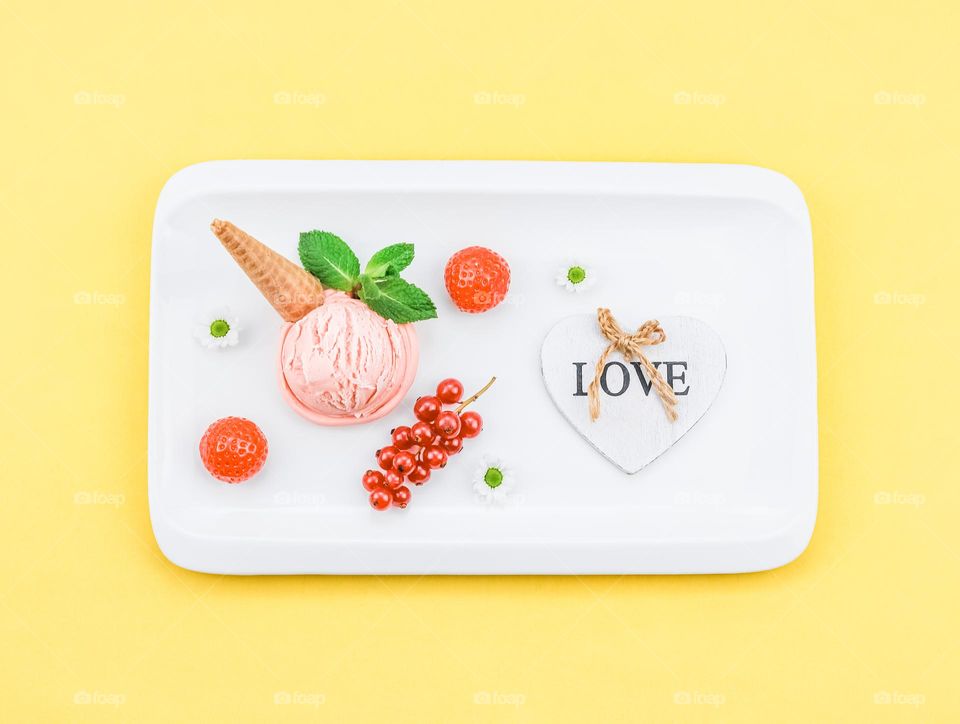 Strawberry ice cream with currants, waffles, rye and a wooden heart with the word "love" lie in a white rectangular plate on a yellow background, close-up flat lay. The concept of summer treats, summer food, natural ice cream.