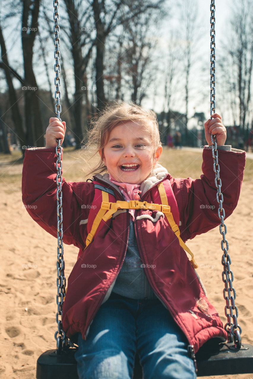 Little smilling happy girl swinging in a park on sunny spring day. Child looking at camera wearing red jacket