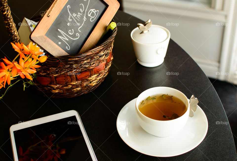 Teatime conceptual photography Beautiful porcelain minimalist style white cup of tea with leaves on table with words "Me Time" handwritten on blackboard in basket with flowers and mobile tablet 
