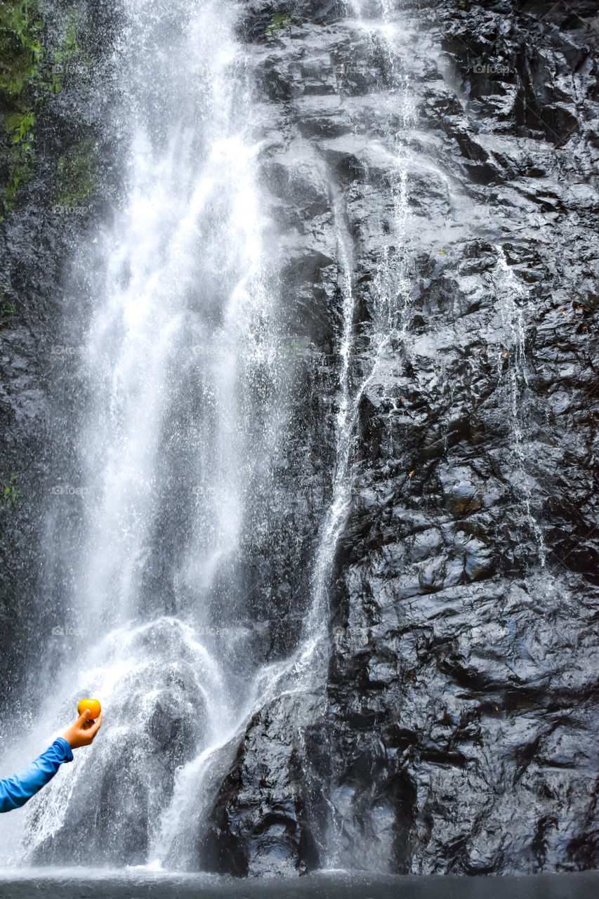 boy wearing blue shirt holding an orange in front of waterfall