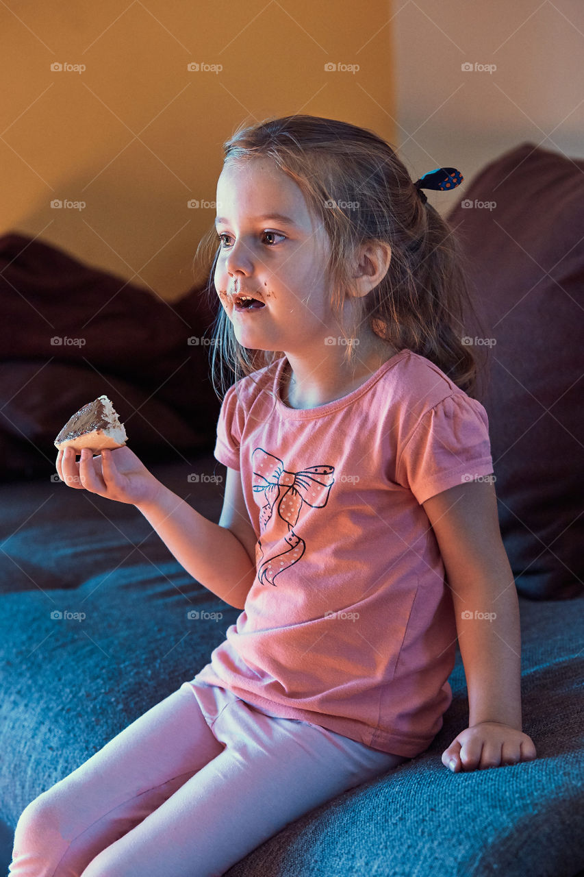 Little cute girl concentrated on watching TV, holding sandwich with chocolate butter, sitting on a sofa in a room. Real people, authentic situations