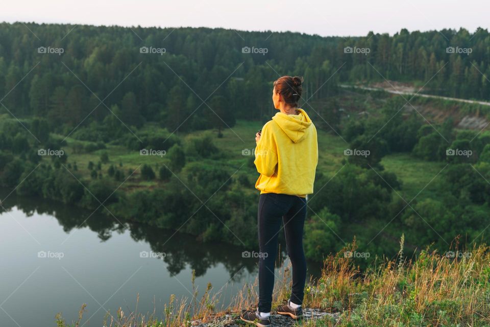 Young woman girl traveler in yellow hoodie with cup of morning coffee looking at sunrise on beautiful view. Local tourism, weekend trip