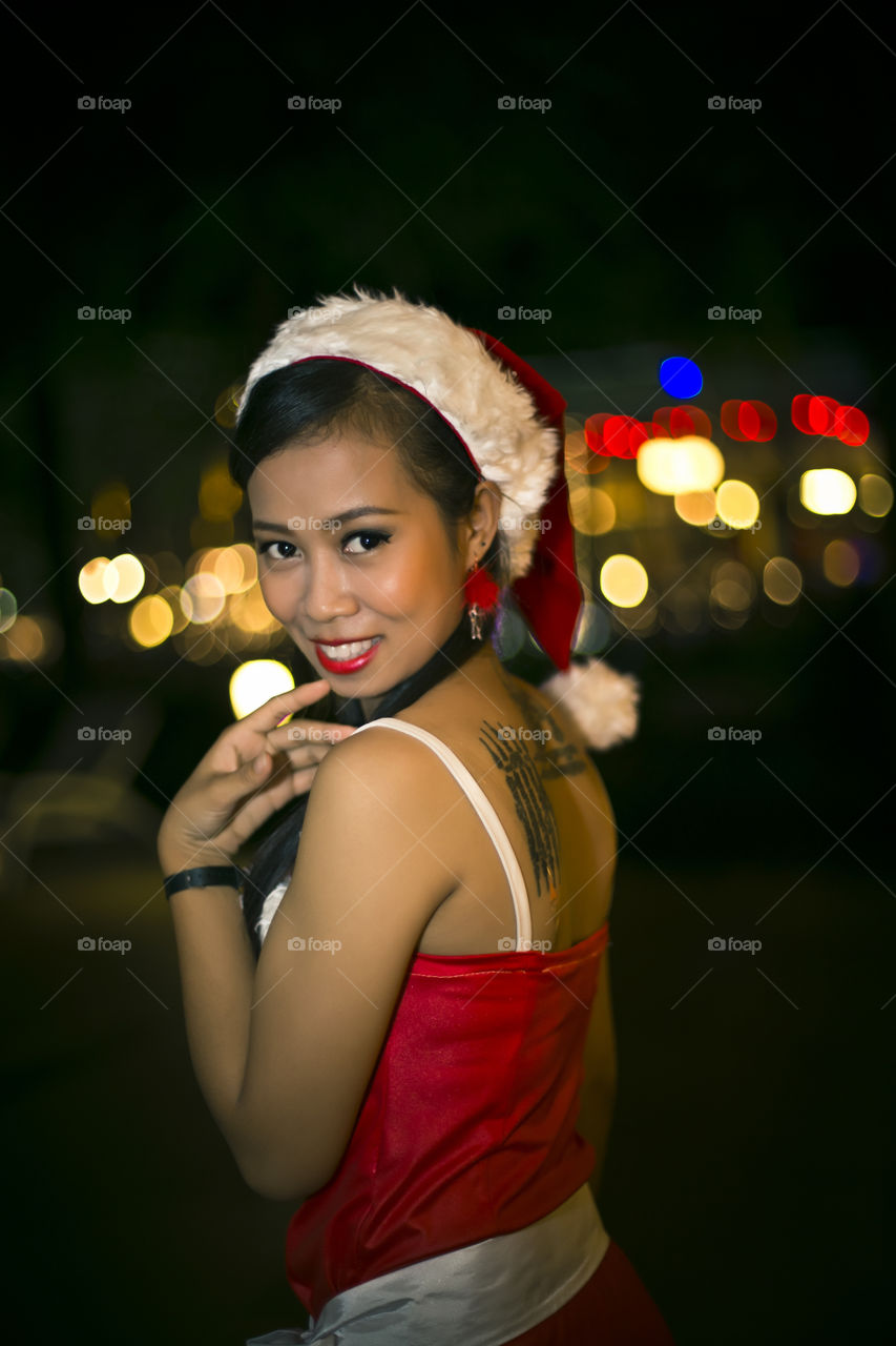 Portrait of pretty Asian Thai girl wearing sexy santaclaus outfit and hat for Christmas party theme celebration at night on a fairy lights bokeh background looking at camera