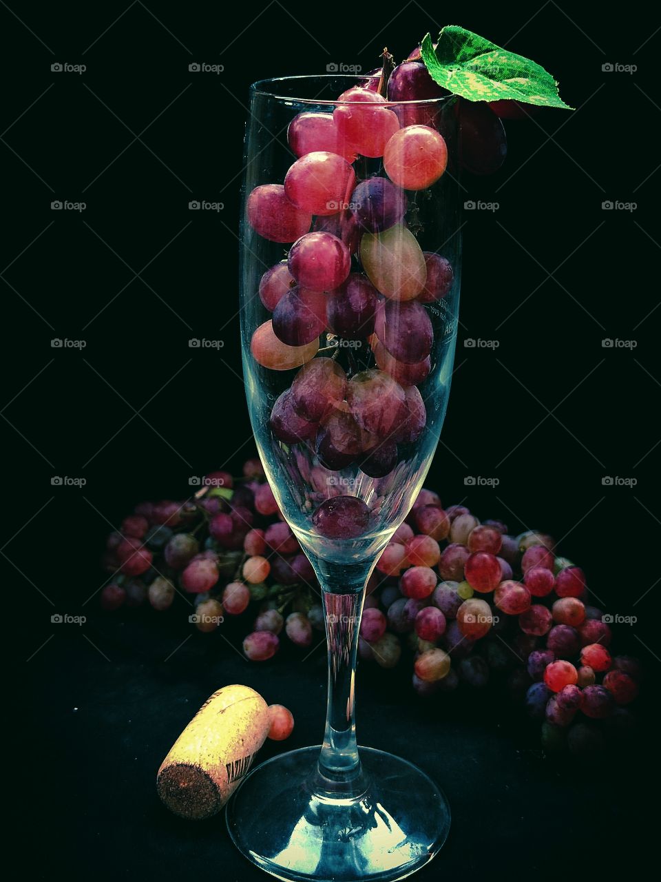 Violet composition.  Glass with purple grapes and green leaves.  Next to the glass is a bunch of purple grapes and a wooden cork from a bottle of wine.