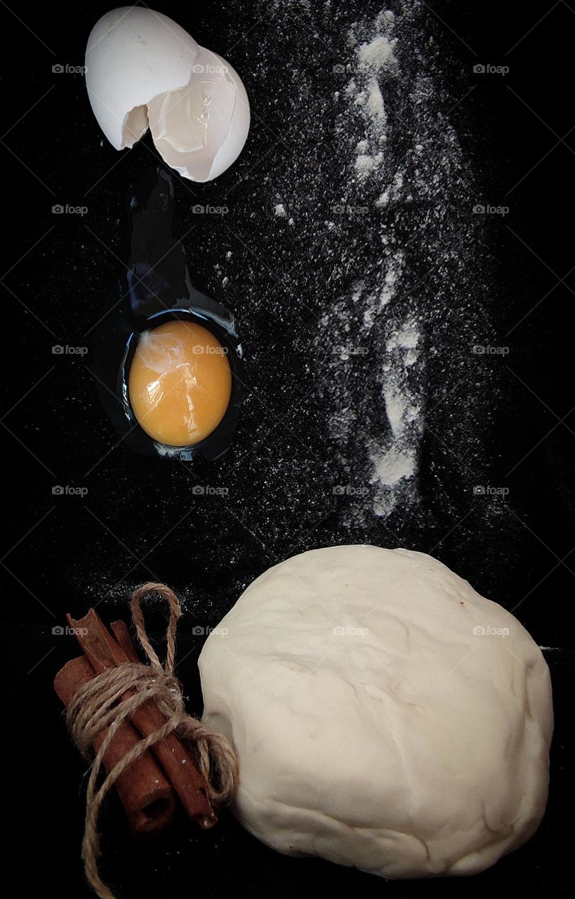 On a black background lies a lump of dough and cinnamon sticks tied with a gray rope.  White flour and a broken egg are poured onto the dough (dough ingredients)