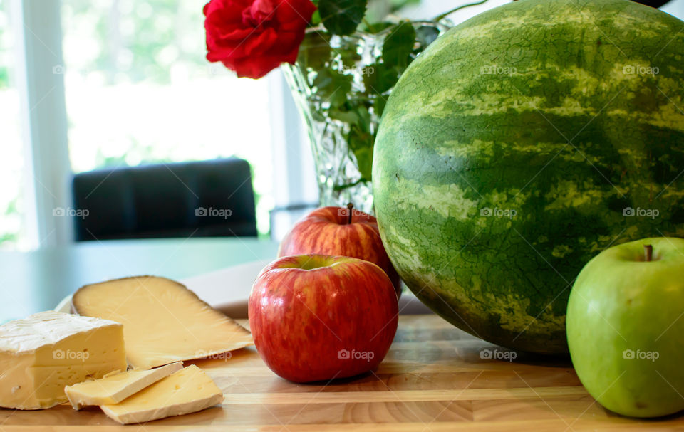 Variety of apples at home on kitchen island on wood with healthy assortment of cheeses and watermelon with flowers and chair in background conceptual healthy food choices and snack photography