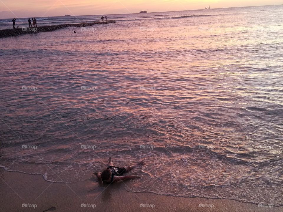 boy lays on the surf line as the sun begins to set over Waikiki Beach