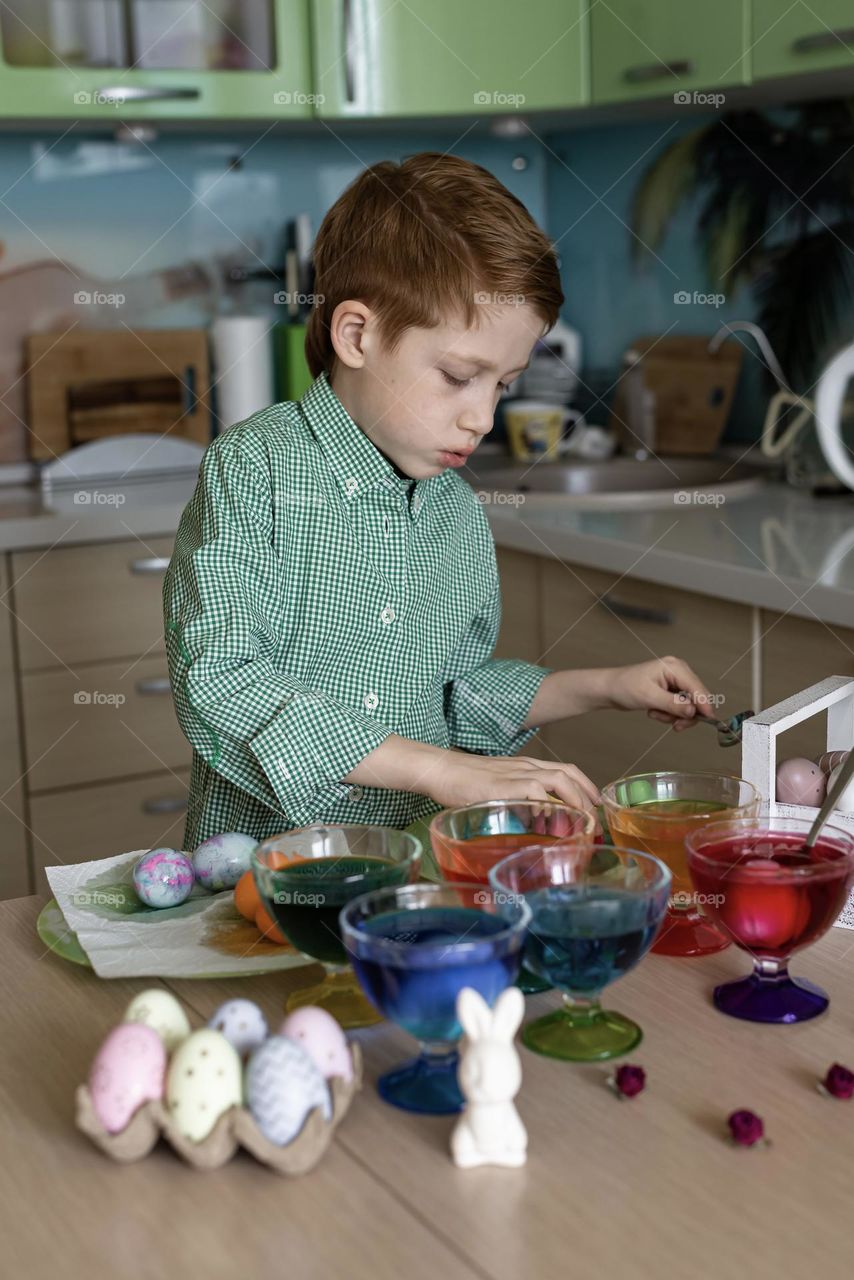 Child red-haired boy in a green shirt paints Easter eggs at home in the kitchen, craft product
