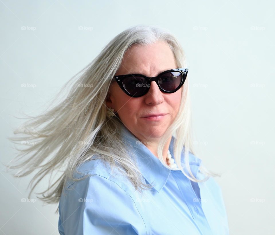 Confident woman with platinum hair, wearing sunglasses showing movement of hair, studio portrait, blue dress shirt, positive attitude, and maturity