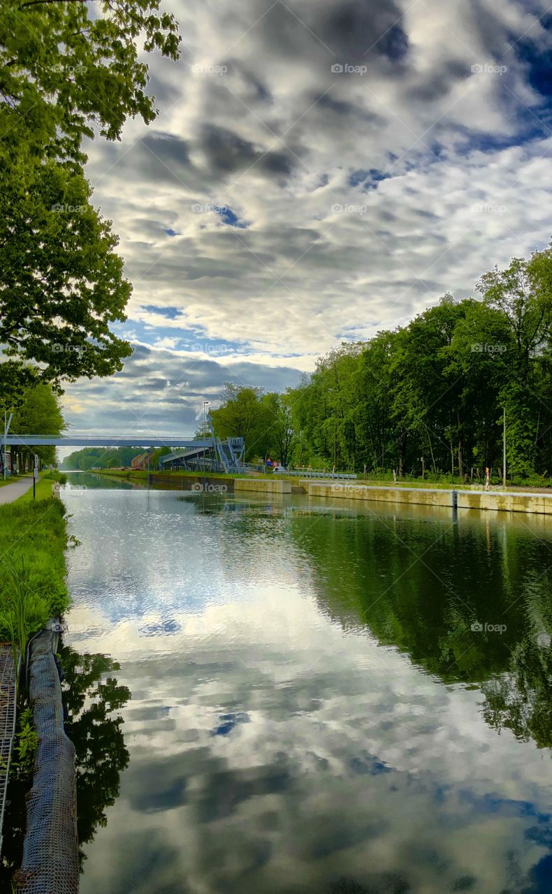 Fluffy white and grey clouds in a blue sky reflected in the water of a canal or river flowing through a rural Countryside forest landscape 