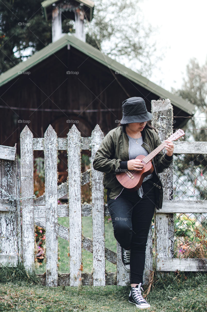 Girl playing the ukulele in front of a fence in the countryside in the middle of nature