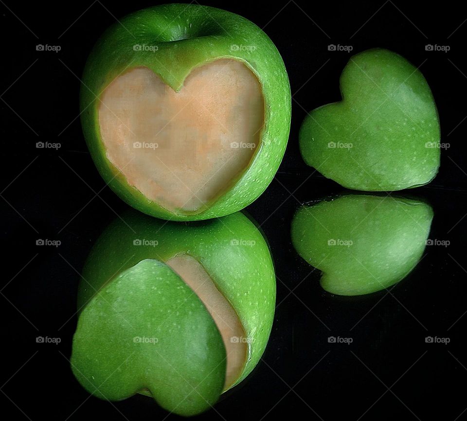 Green color. On a black mirror background lies a green apple from the surface of which a part in the form of a heart is cut off. Nearby is a green heart made of an apple. In the reflection is a green apple with a piece of a green apple heart