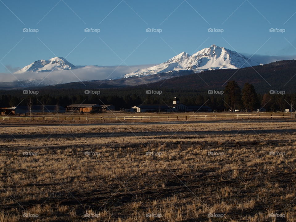 The majestic snow covered Three Sisters in Central Oregon’s Cascade Mountain Range overlook a farm field during the off-season on a beautiful sunny day.