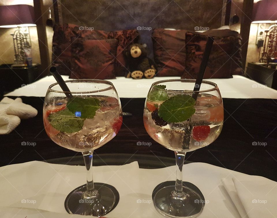Gin and Tonic drinks with berries and mint leaves with ice and straws in glasses beside bed with pillows and teddy bear