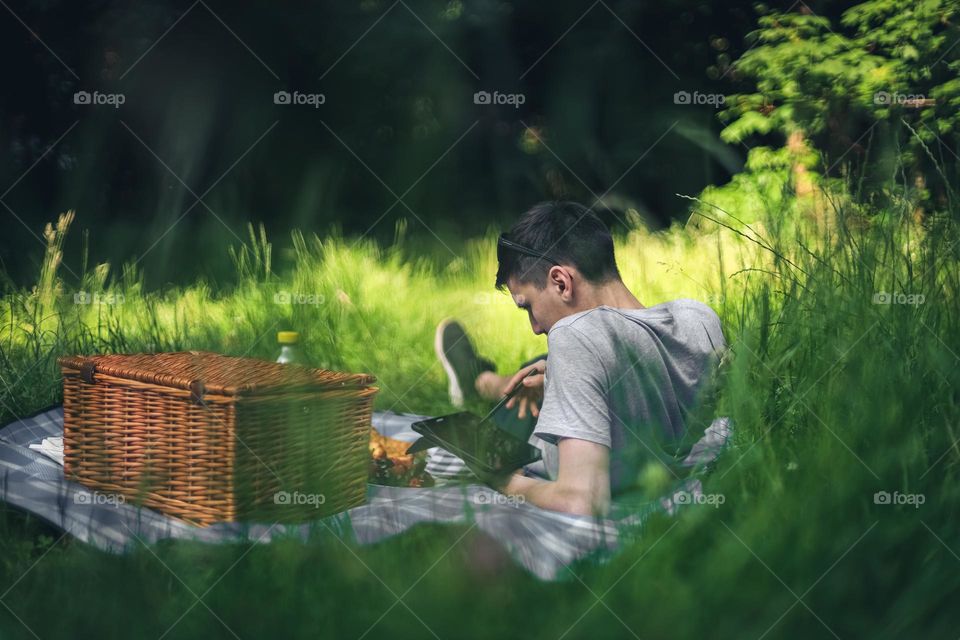 One young handsome caucasian guy lies on a bedspread with a wicker basket, spoiled food, drinks and plays on a tablet on a green meadow with tall grass on a summer sunny day, close-up view from below.