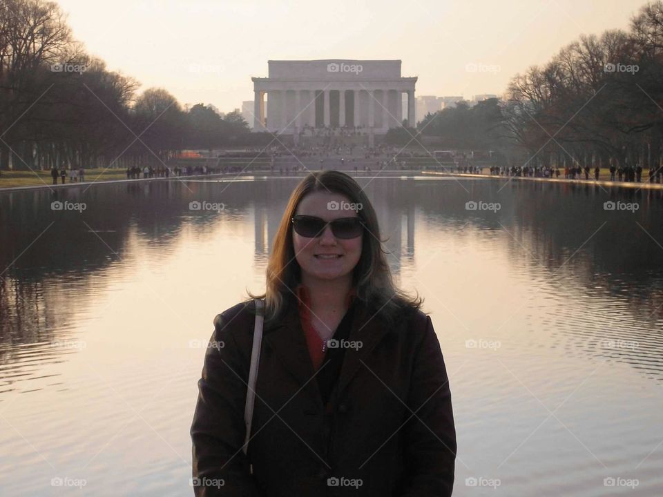 A woman stands wearing sunglasses and a leather coat with the National Mall and Lincoln Memorial behind her. The Lincoln Memorial is located at the western end of the National Mall in Washington DC. The trees lining the water reflect in the pool. 