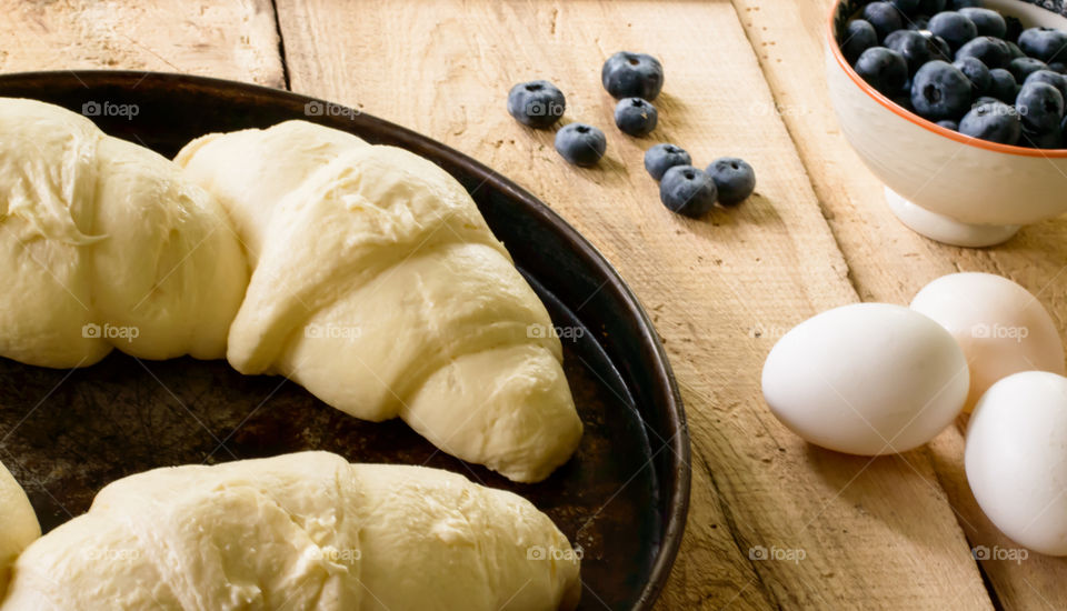Baking fresh bread - croissants on baking tray next to fresh eggs and blueberries on rustic wood table conceptual baking photography
