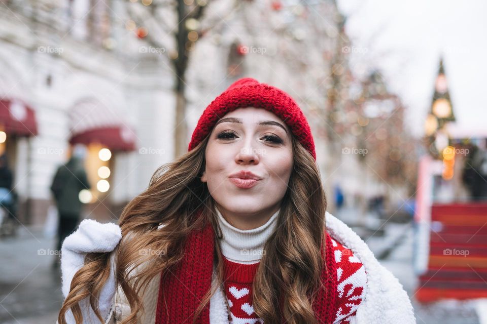 Young happy woman with curly hair in red knitted hat at the Christmas fair in winter street decorated with lights