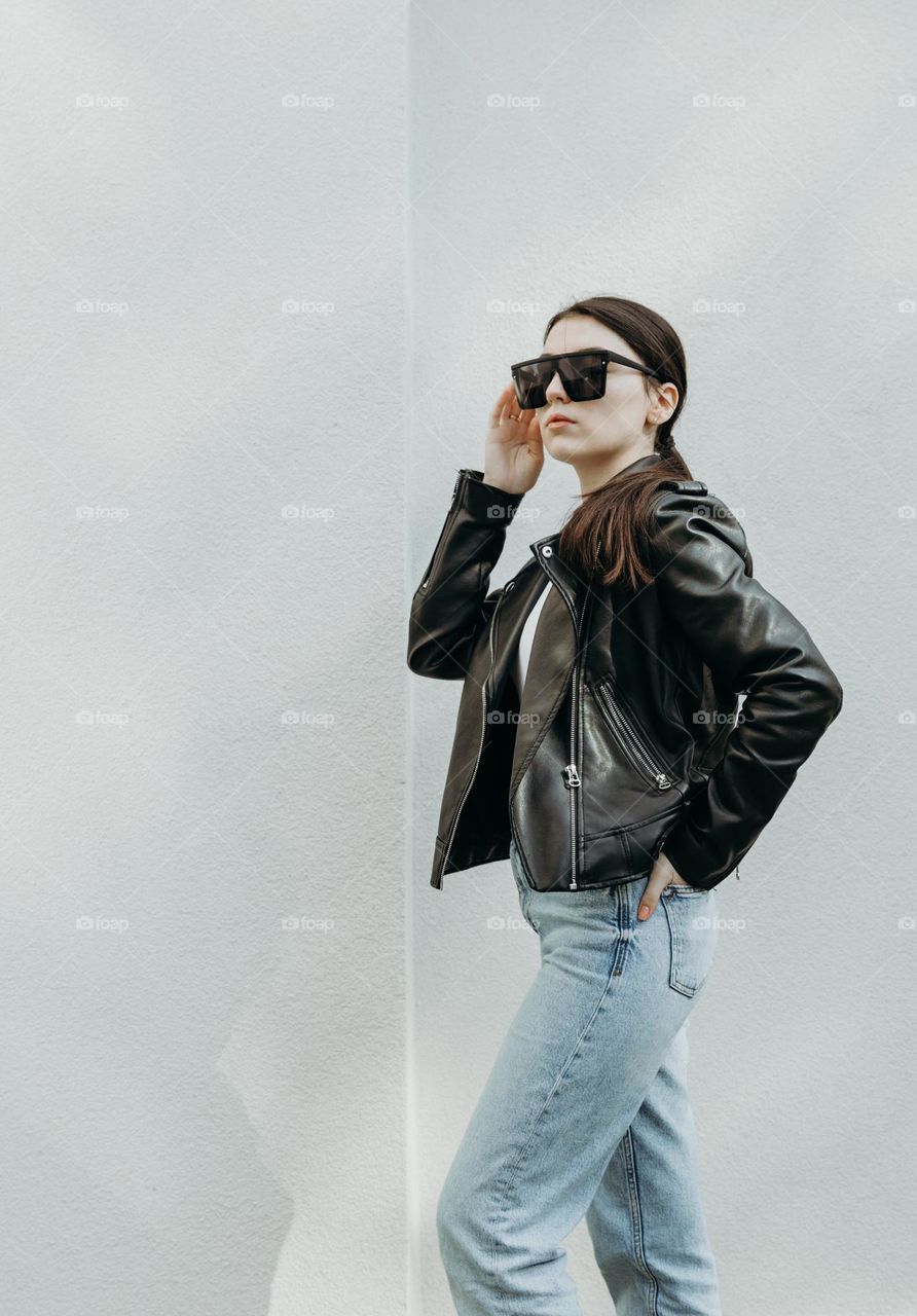 Portrait of one beautiful young Caucasian brunette girl in stylish leather jacket and sunglasses with serious emotion on her face stands sideways against white street wall with small space for text on summer evening, side view close-up
