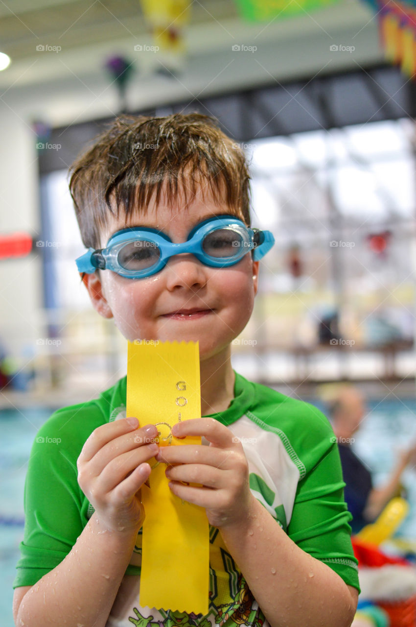 Young toddler boy wearing goggles at an indoor swimming pool while smiling and holding an award ribbon after a swim lesson