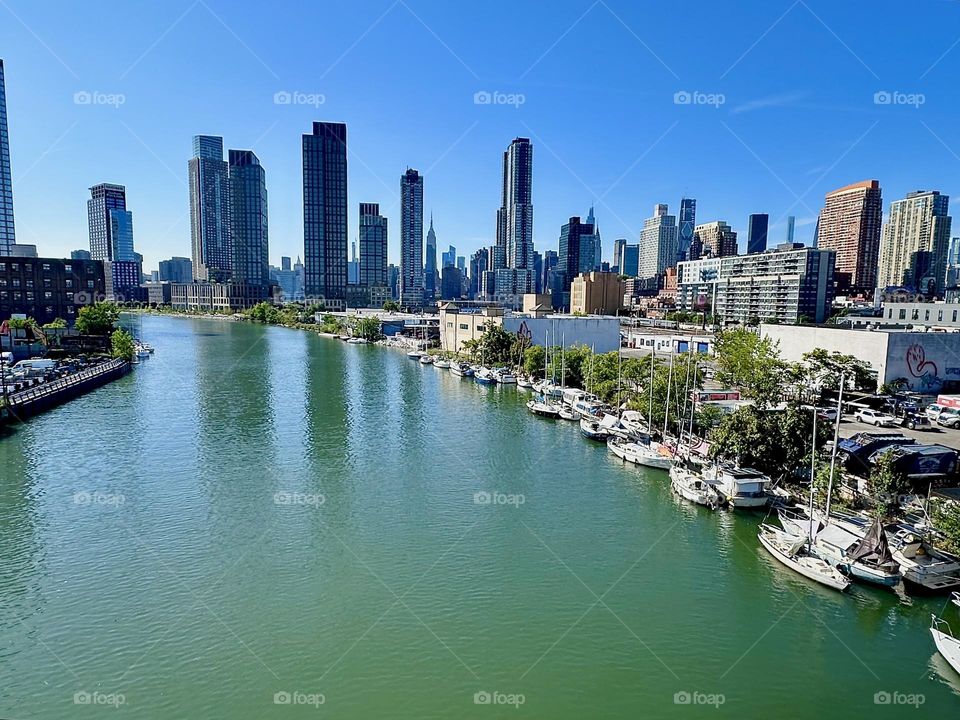 This is picturesque “Newtown Creek” with its variety of boats seen from the “Pulaski Bridge” that connects “Greenpoint”, Bklyn to LIC, Queens. Across the water we see “Manhattan” including the “Empire State Building”. 2024. Hypnotic Productions