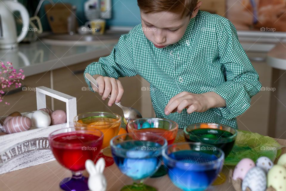 Child red-haired boy in a checkered shirt paints Easter eggs at home in the kitchen. DIY cooking