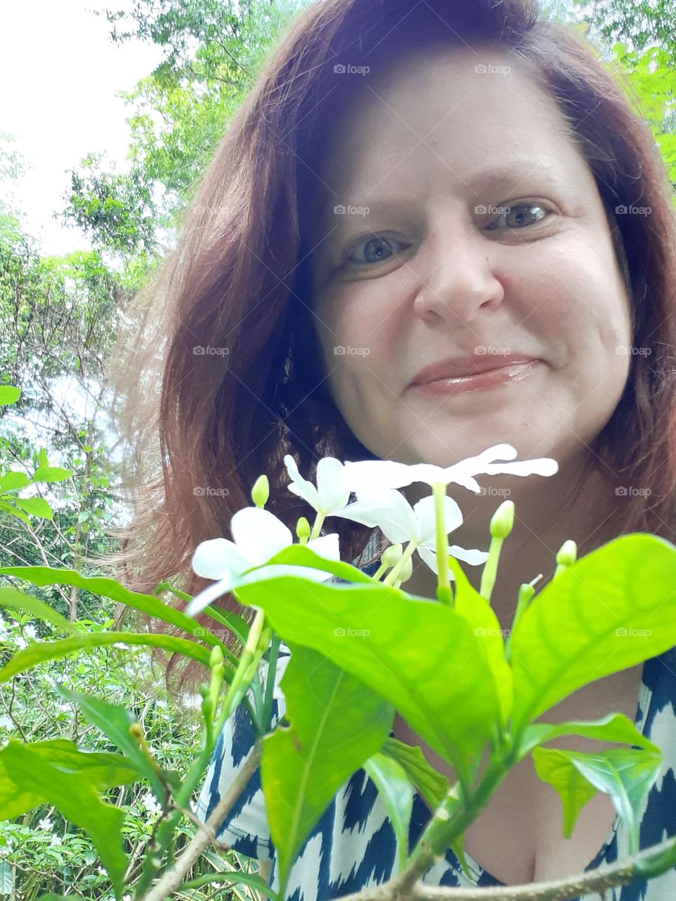 A middle aged caucasian woman is smiling as she stands in a garden and smells the flowers.