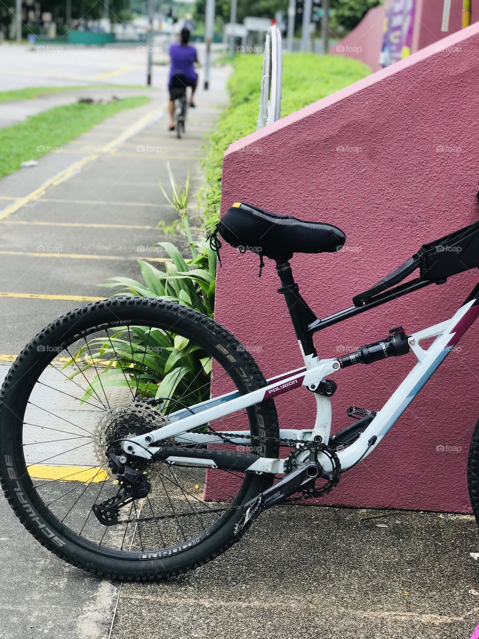 A black colour bicycle stands in front of the pink colour wall at the same time a person going on bicycle at back of the wall 