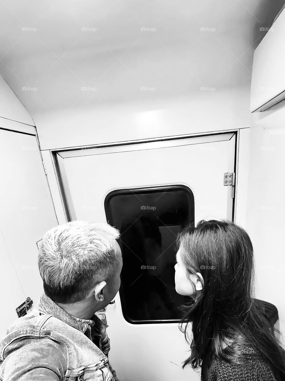 Portrait of a young couple standing face to face in a room that looks cramped. They faced a square window with curved edges. This image is black and white and has high contrast between light and shadow