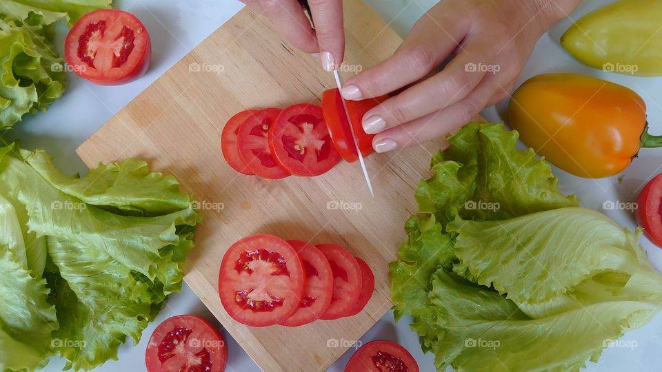 Time to cook! Cutting tomatos. On the kitchen table, a wooden cutting board, a ceramic knife, tomatoes, sweet peppers, lettuce