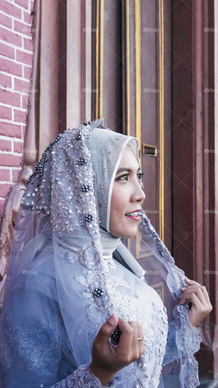 Portrait of young and beautiful Asian muslim woman wearing silver robe with matching hijab and headscarf, holding headscarf and smiling, Red brick wall background. for the wedding.