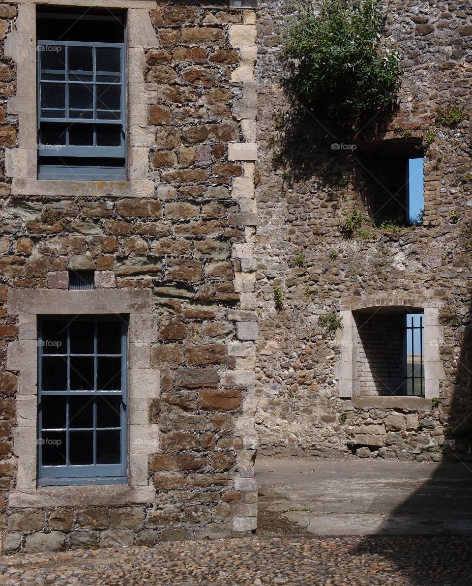 Staggered stone walls in an English courtyard with empty windows with bars and glass windows in blue frames on a sunny summer day. 
