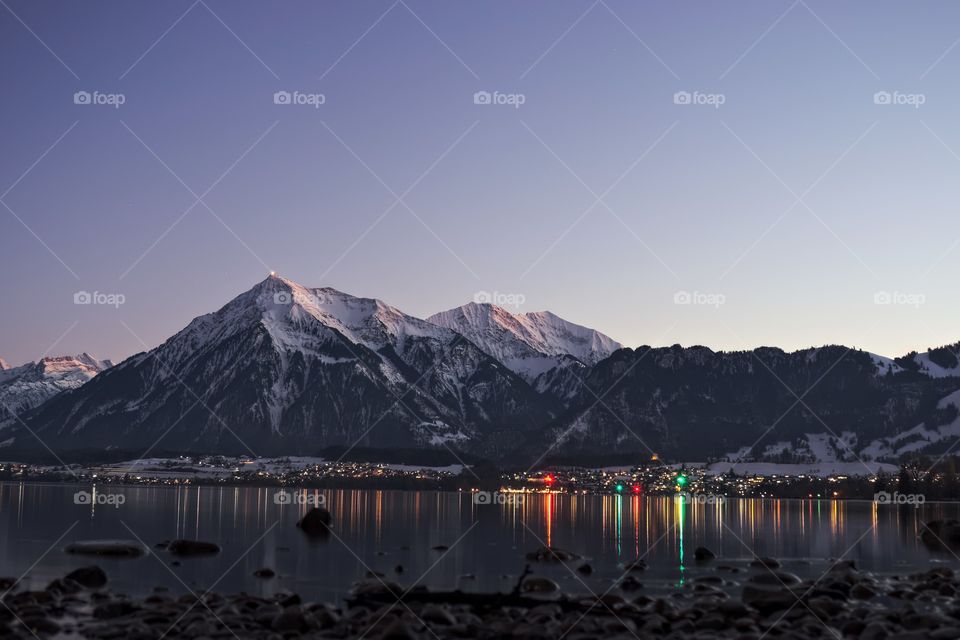 Snow capped mountain above village with lights reflected in lake.