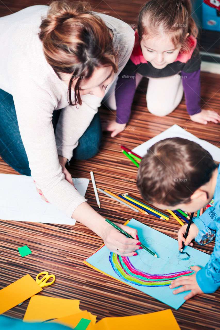 Mom helping her little son drawing a colorful picture of car using pencil crayons sitting on floor. Shot from above